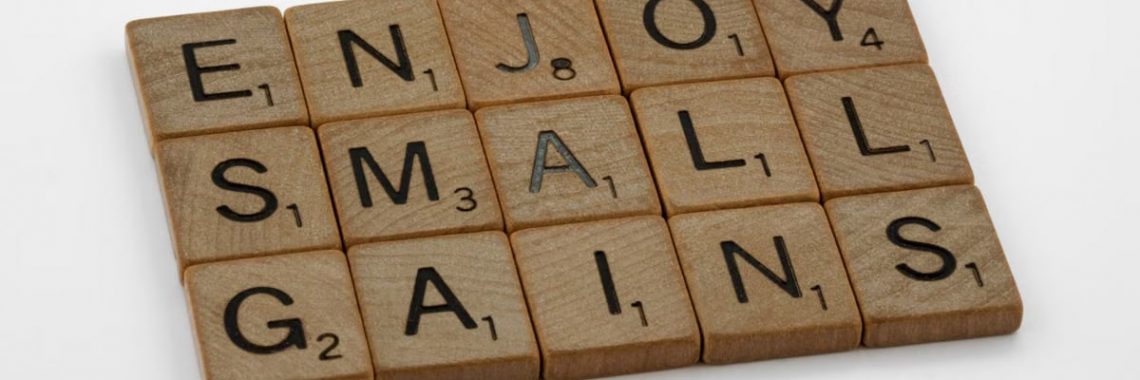 Close-up of Scrabble tiles spelling 'Enjoy Small Gains' on a white surface.